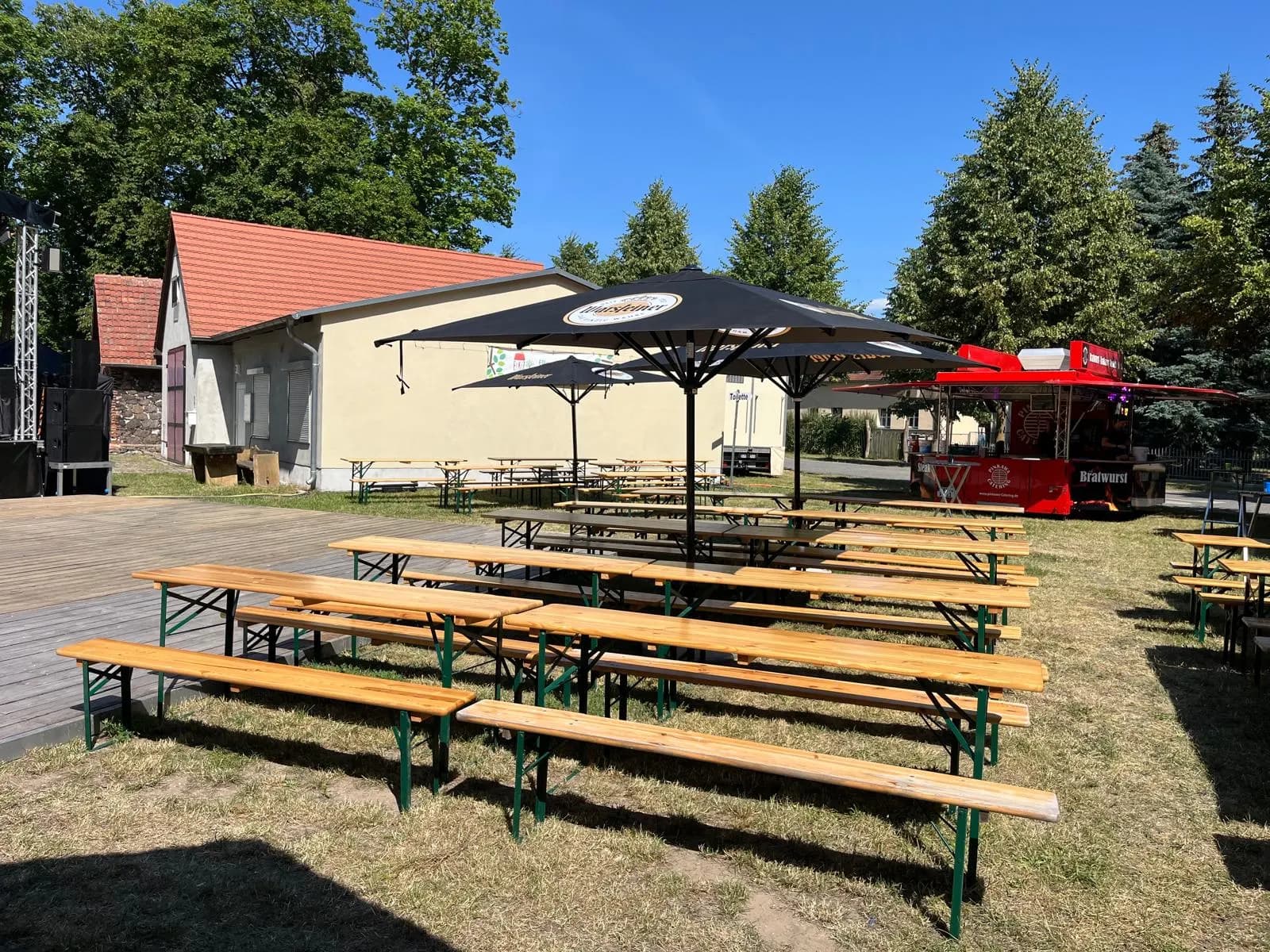 Aufgereihte Bierzeltgarnituren mit Sonnenschirm, im Hintergrund der rote Grillwagen