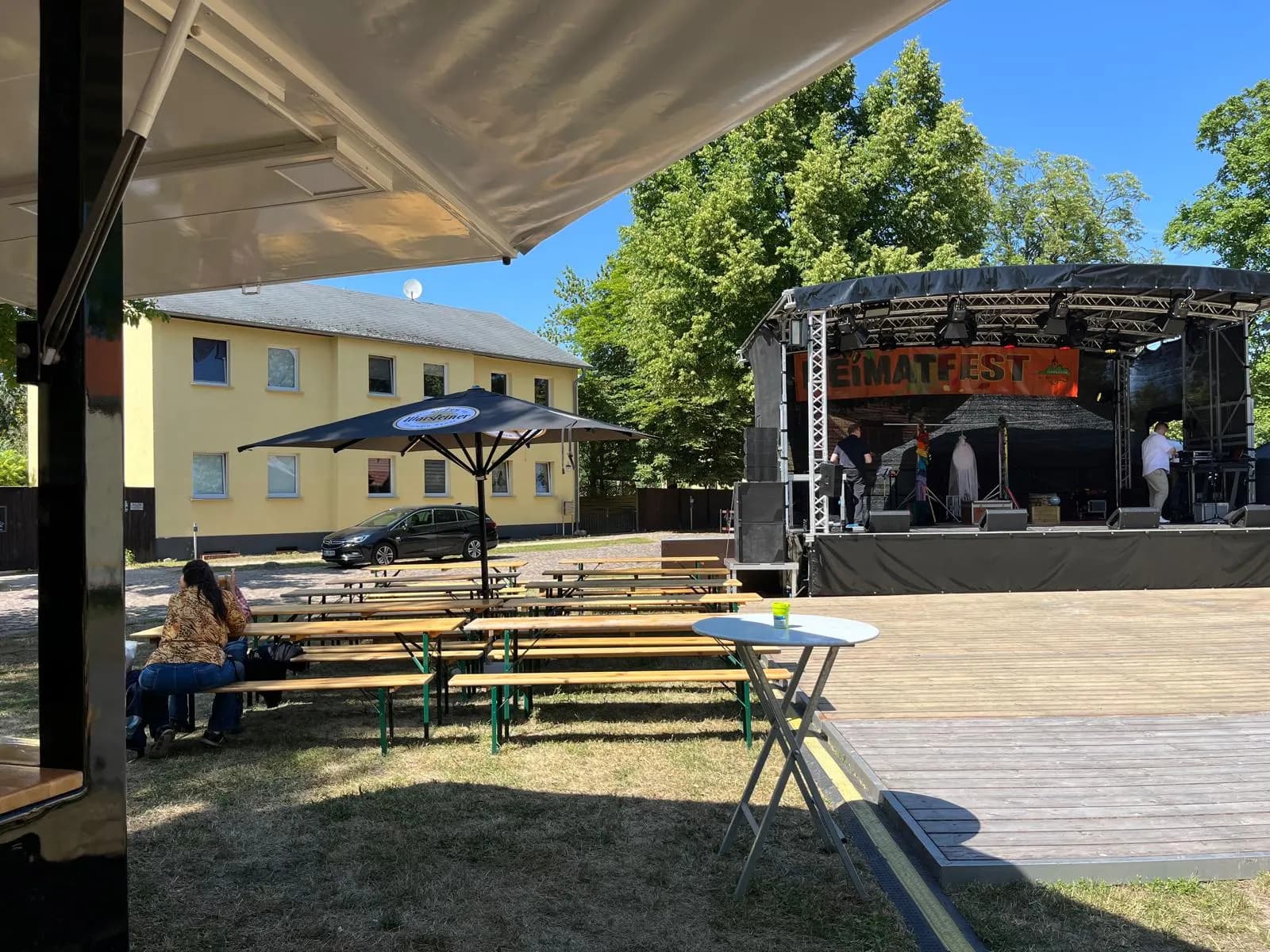 Festgelände mit Bühne, Biergarnituren und Sonnenschirmen an einem sonnigen Tag