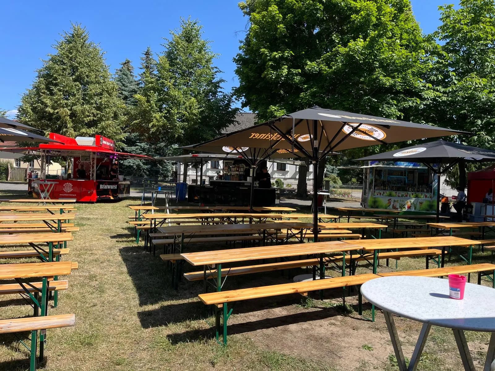Veranstaltungsgelände mit langen Tischreihen unter Sonnenschirmen, im Hintergrund der rote Grillwagen