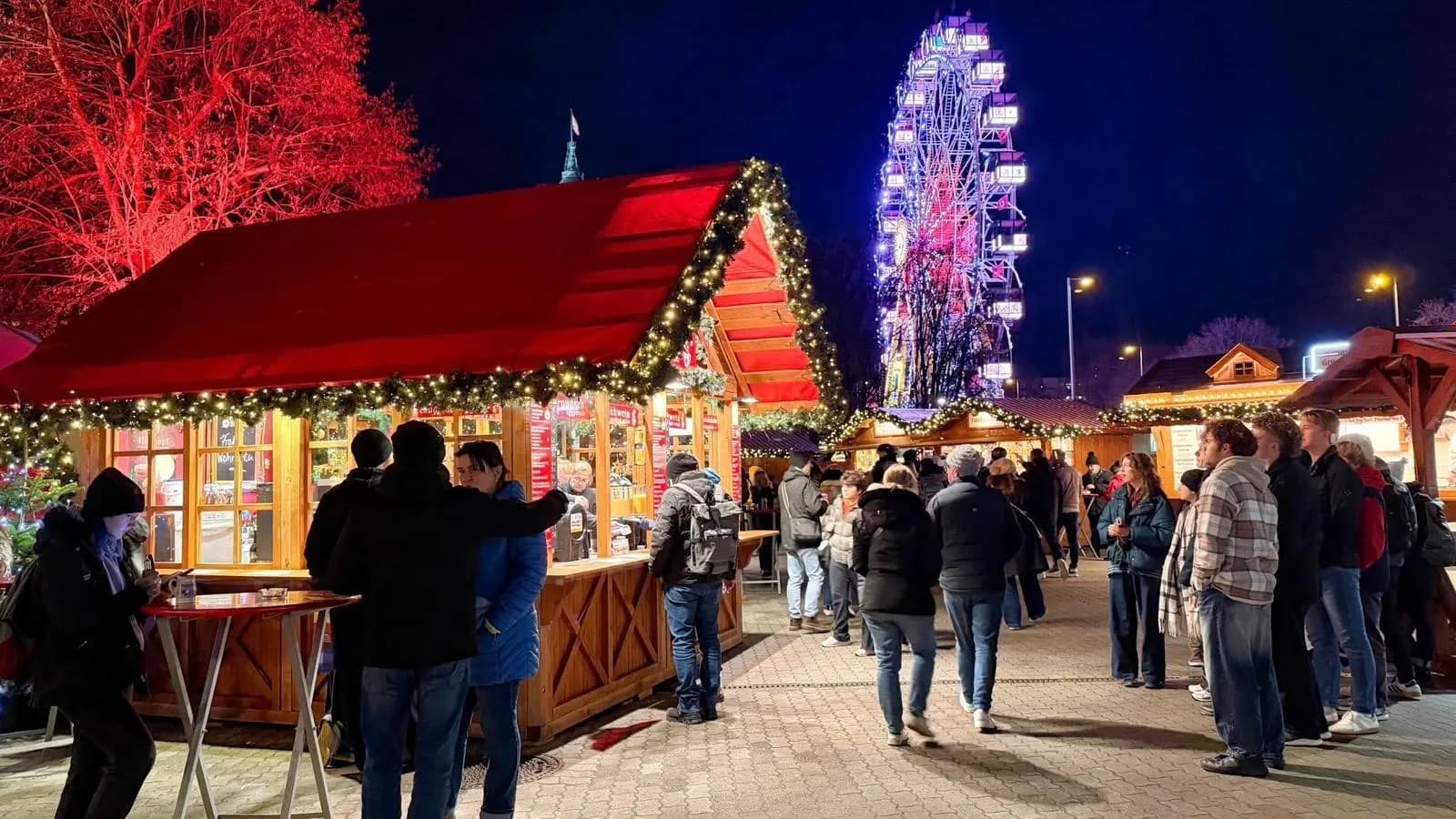 Weihnachtsmarkt bei Nacht, im Hintergrund ein beleuchtetes Riesenrad
